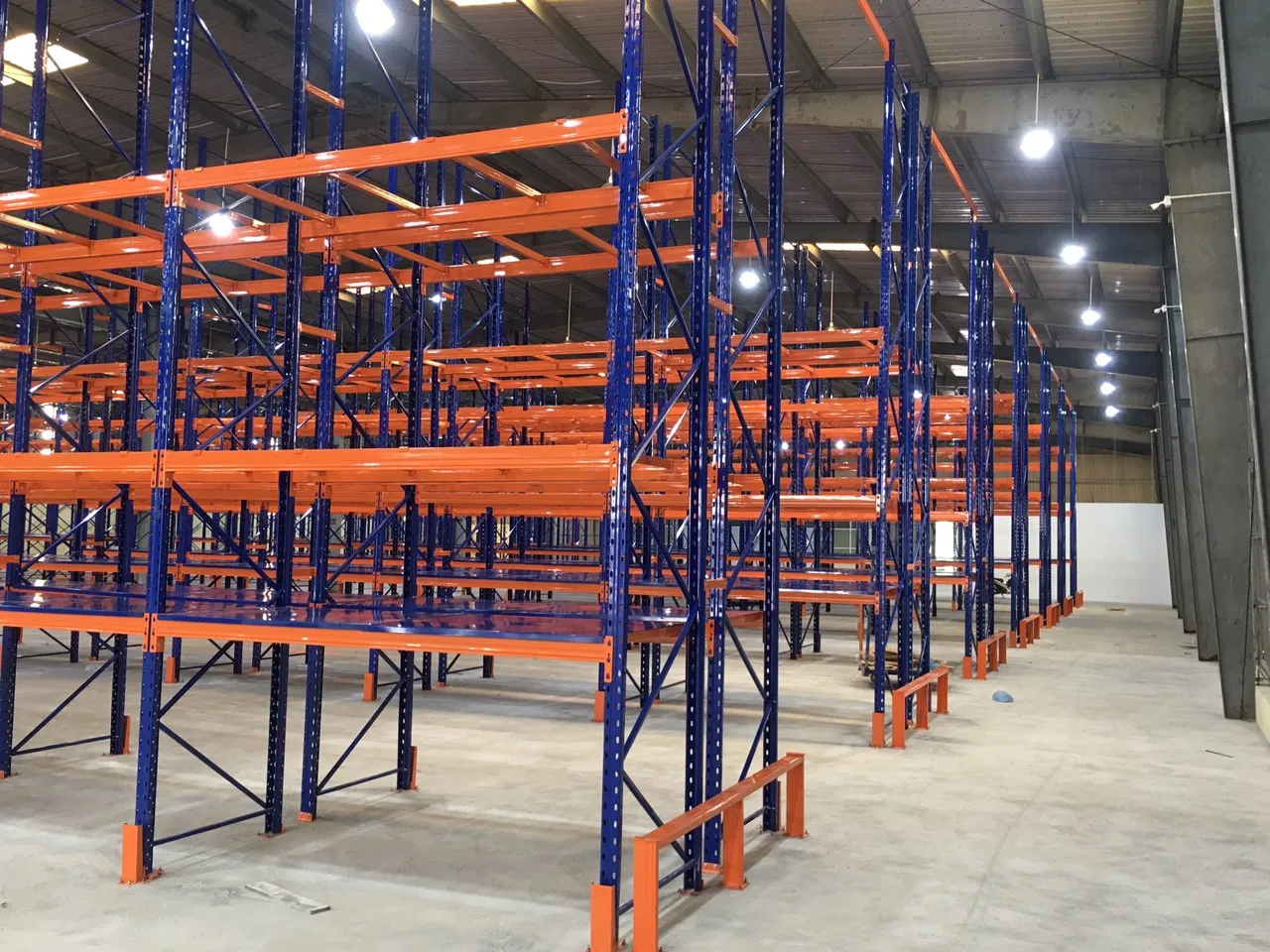 Nhiều dãy kệ pallet cao tầng trong kho có đèn chiếu sáng treo trên mái